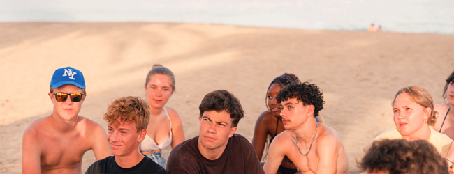 Mehrere Jugendliche sitzen im Sand am Strand von Barcelona und geniessen die Abendsonne